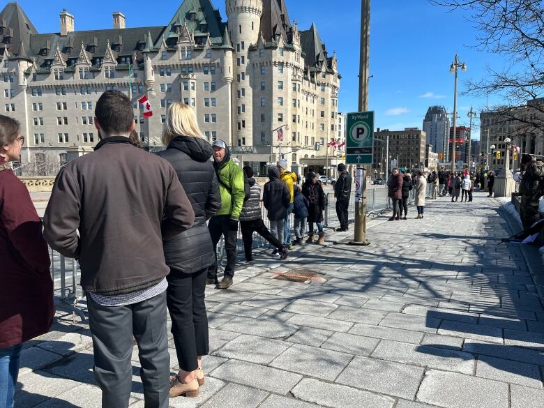 A single-file line of people along a temporary fence that blocks a road.