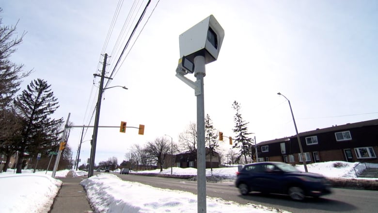 A dark-coloured car passes by an automated speed camera beside a four-lane road in winter