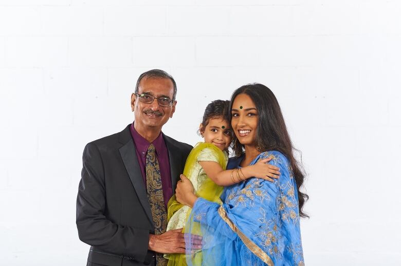 A older man, his young grand-daughter and daughter pose in front of a white backdrop for a family photo. 