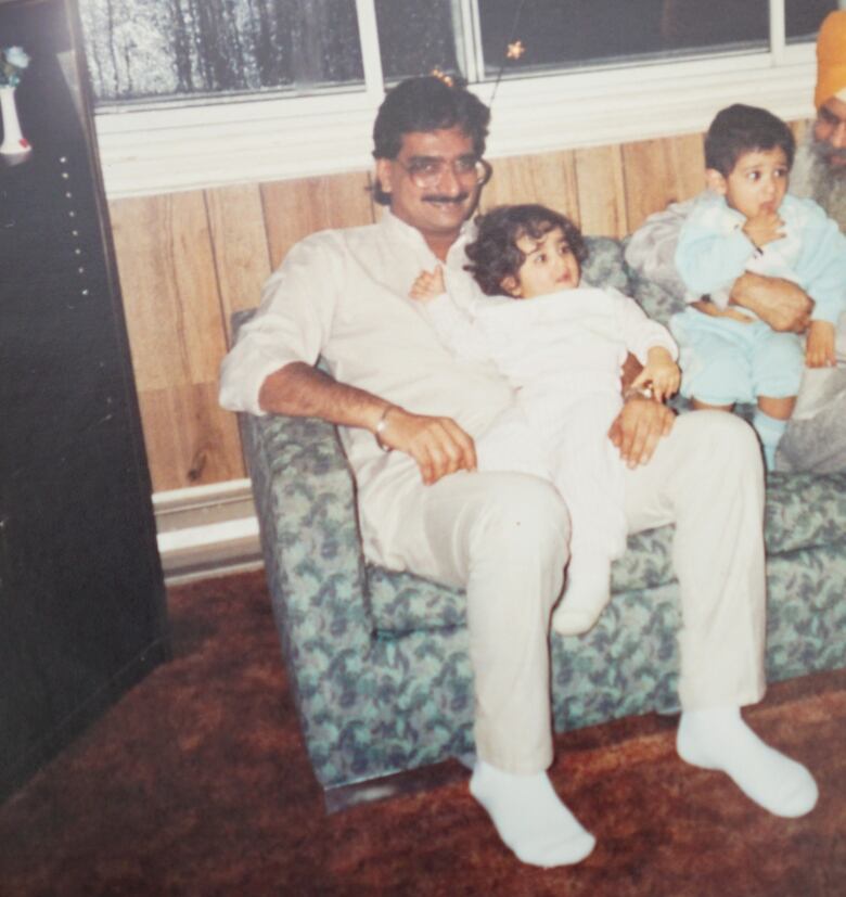 A smiling man dressed in white sits on a couch with his toddler daughter. 