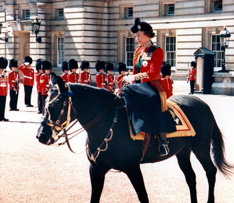 A person rides a horse sidesaddle in front of soldiers standing in formation.