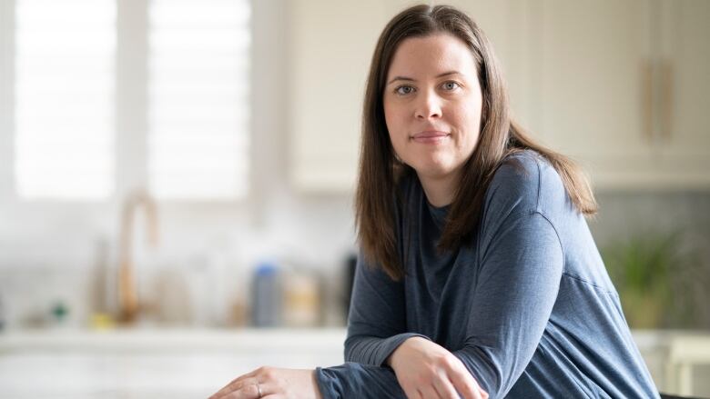 A smiling woman with long brown hair and wearing a blue shirt leans against a marble counter.