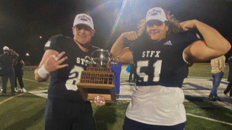 Two football players pose with a championship trophy on the sidelines of the field.