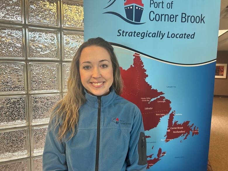 A woman with long, blonde hair stands in front of a banner for the Port of Corner Brook. She's also wearing a blue zip-up sweater with the corporation's logo on it.