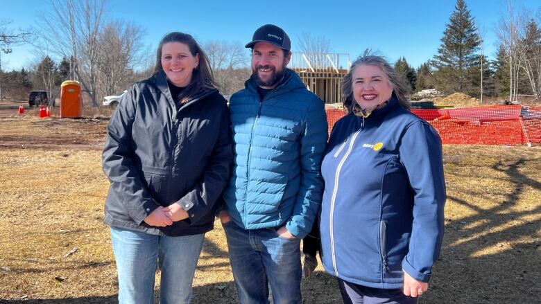 A woman wearing a dark coat, a man wearing a blue coat and a woman wearing a blue coat stand in front of a construction site on a sunny day.