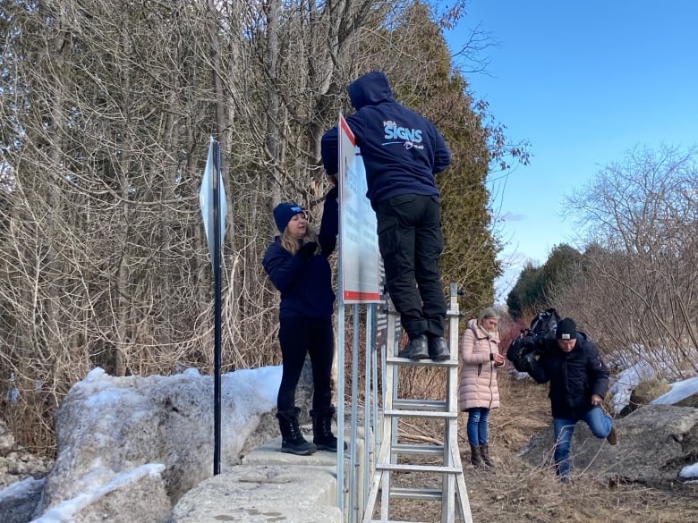 Two workers in blue hoodies install a sign in a forested area while a media crew stands in the background.