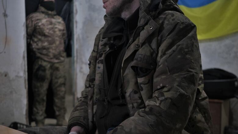 A person in military garb is seen seated as another guards the doorway.