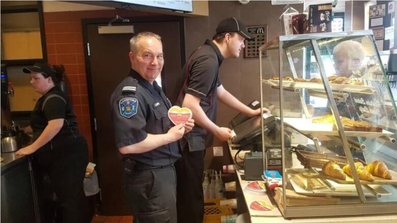 Assistant Chief Lance Caven stands in front of a display case of baked goods holding a paper heart.