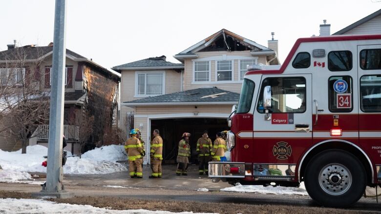 Firefighters stand in a driveway outside a home with a fire-damaged roof. A fire truck is parked out front. The home to left has burned siding.