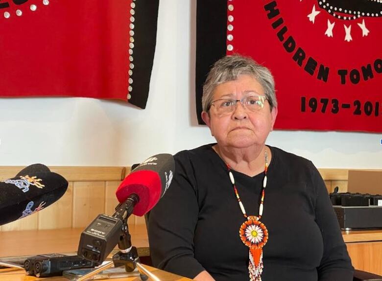 A woman wearing a beaded necklace sits at a table with microphones.