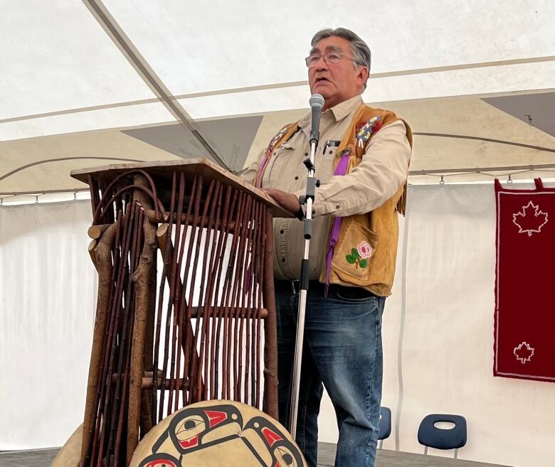 A man stands speaking at a podium on stage, with two Indigenous drums leaning against the podium.