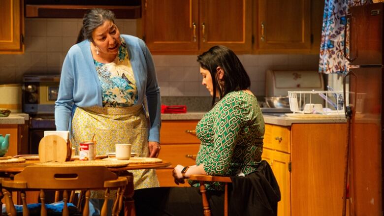 A scene from a play shows an older woman standing in a kitchen set, speaking to a younger woman sitting at a table. The younger woman is looking down.