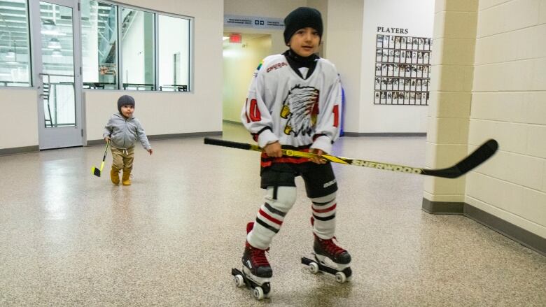 Two boys carrying hockey sticks run around a hallway.