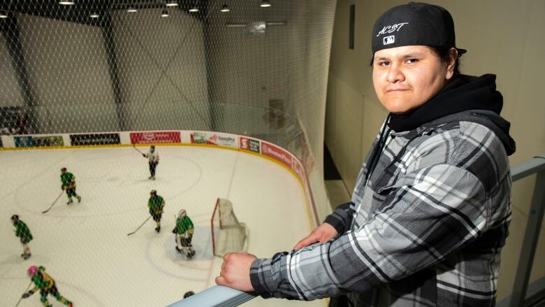 A young man wearing a ball cap stands in front of a ice rink with hockey players.