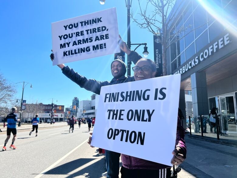 A man and a woman cheering on passing runners. Each of them is holding a sign, one of them says 