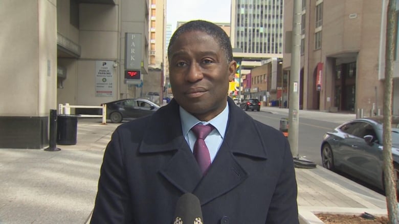 A formally dressed man stands in front of a microphone on a city sidewalk.