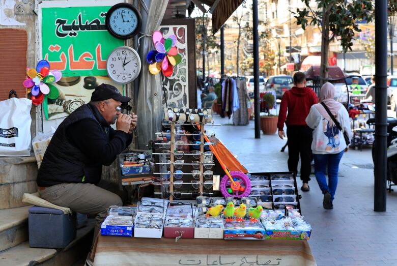 A man sits at a table, covered in various items, along a sidewalk. Two clocks showing different times hang above his head. 