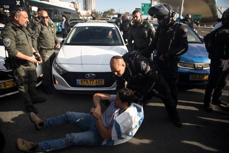 A man sits on the pavement in front of a vehicle as another man attempts to move him.