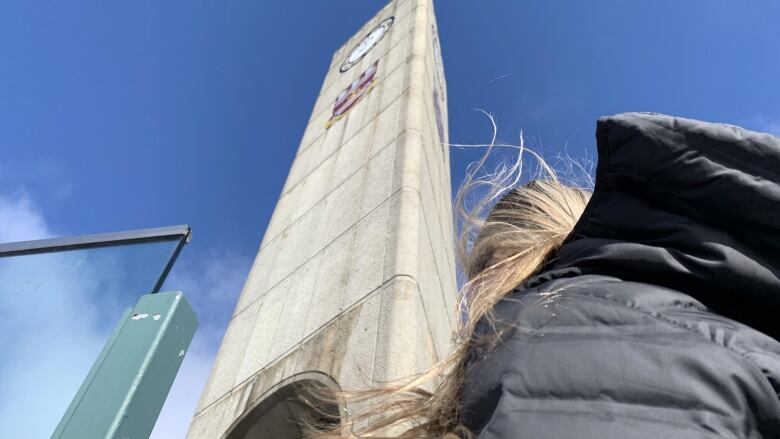 A person with blonde hair and wearing a black jacket is back on, and is looking up at a clock tower. 