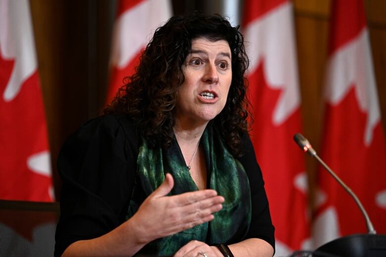 A woman speaks at a microphone in front of a row of Canadian flags.