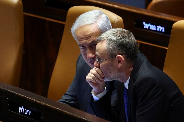 Two men sit next to each other in brown leather seats inside a chamber room. One leans forward, with his hand covering his mouth.