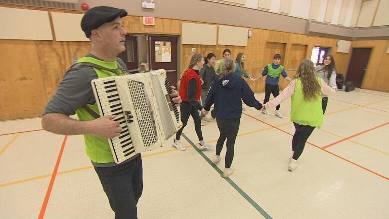 A man plays an accordion while students dance in a circle in the background.