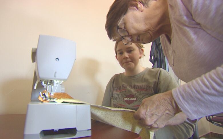 An older woman looks over a young woman's shoulder and shows her how to use a sewing machine.