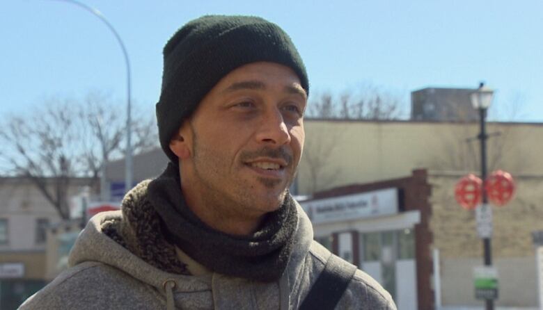 Young man wearing a dark touque and warm jacket standing on a city street with a slight smile.