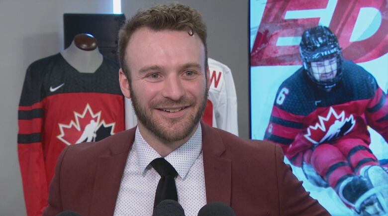 A male para ice hockey player stands behind reporter microphones with photos of himself playing the game in the background. 