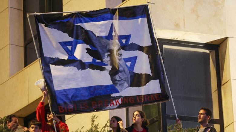 A group of young protesters, standing on a balcony lined with shrubs, lift a large flag depicting a ripped flag of Israel and the face of a man. Written underneath are the words: Bibi is breaking Israel.