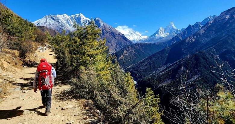 A man in a red backpack walks along a dirt path up the side of a mountain, with snow-covered peaks in the distance. 