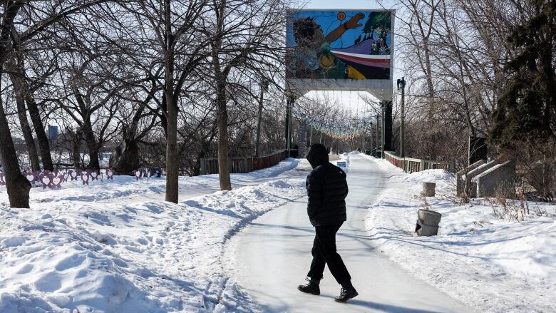 The black silhouette of a person walking across a snowy ground, a bridge in the background.