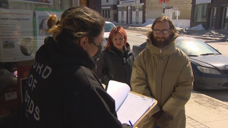 A woman with a clipboard stands on the step of a food bank building and speaks to man in a beige winter coat with long hair and glasses.