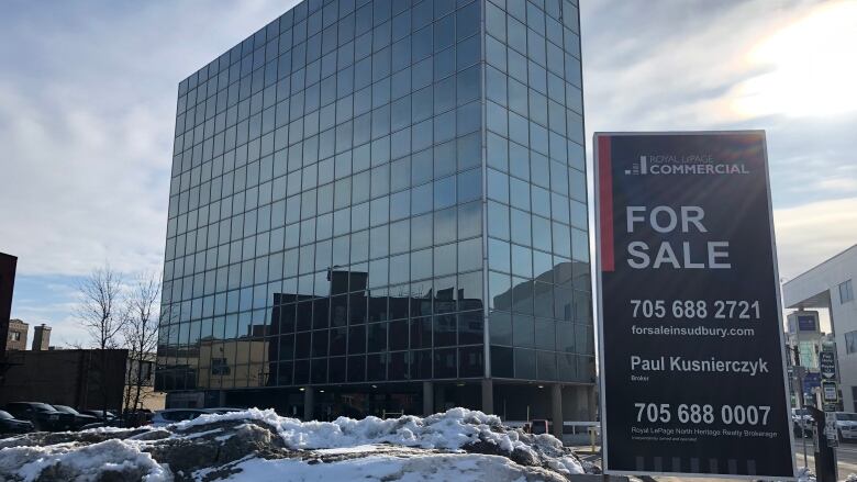 A for sale sign stands in front of a 16-story high-rise building with mirrored windows.