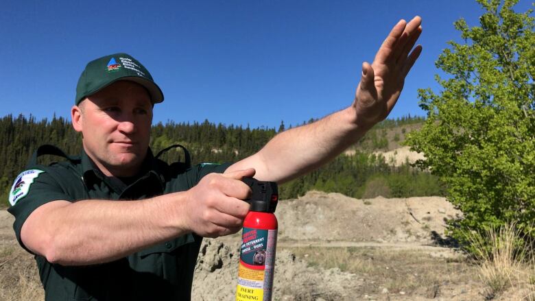 A uniformed conservation officer holds a cannister of bear spray in front of him while also partially shielding his face in a use demonstration.