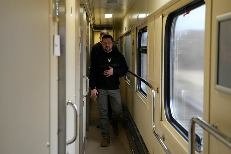 A man in a black sweatshirt and grey pants walks down the aisle of a train.