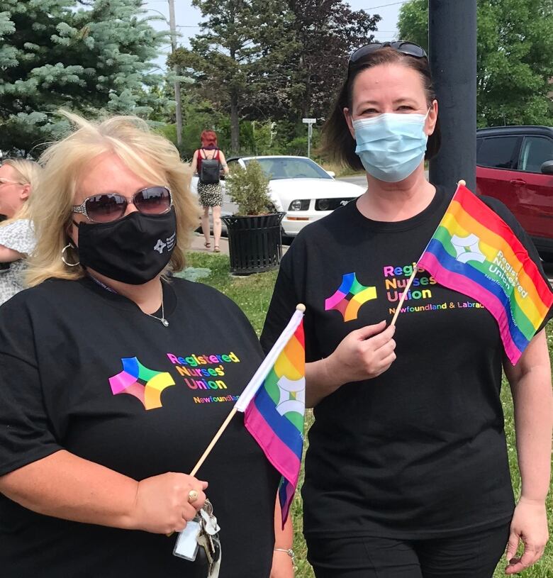 Two women are standing next to each other. Both wear face masks and T-shirts that read Registered Nurses' Union, with the letters printed in multiple colours. Both are holding a pride flag each in their hands.