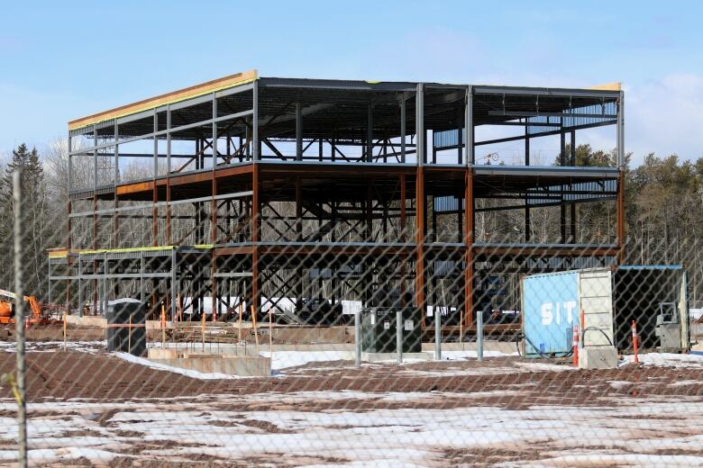 A steel frame of a three-storey building rising over a construction site with concrete footings and machinery.  