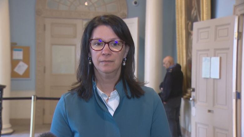 A woman with shoulder length brown hair and  glasses wears a teal blazer. She is standing between two pillars.