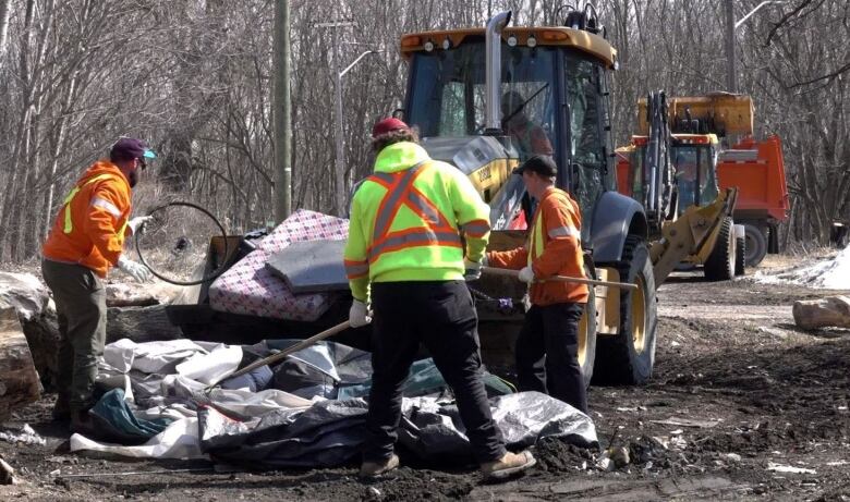 Four men in fluorescent jackets use pitchforks to shove a green and grey tent into the bucket of a front-end loader. 