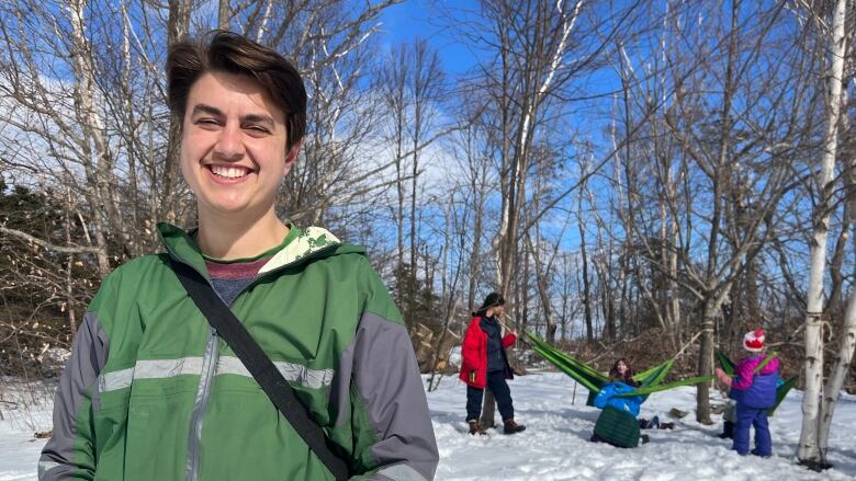 A short haired person smiles at the camera while bundled up kids play in the snow in the background