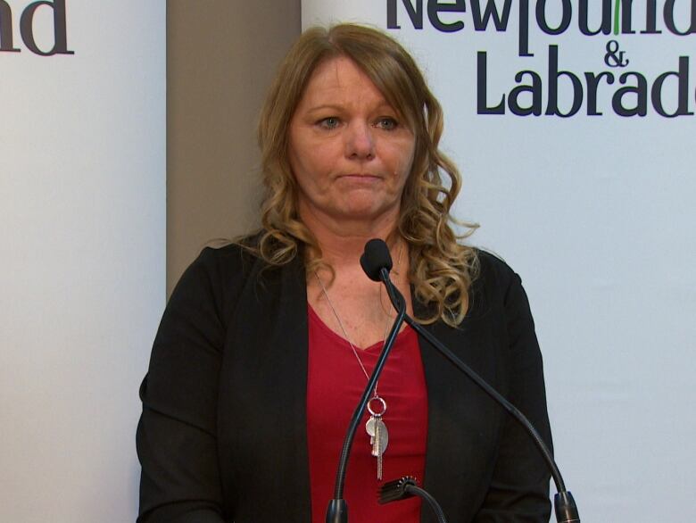 A woman wearing a red shirt and a black jacket stands in front of a pair of microphones.