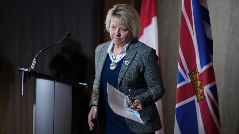 A woman with a blond bob haircut, wearing a grey blazer over a navy blue sweater steps away from a podium. In her left hand, she holds speaking notes and a pair of reading glasses.