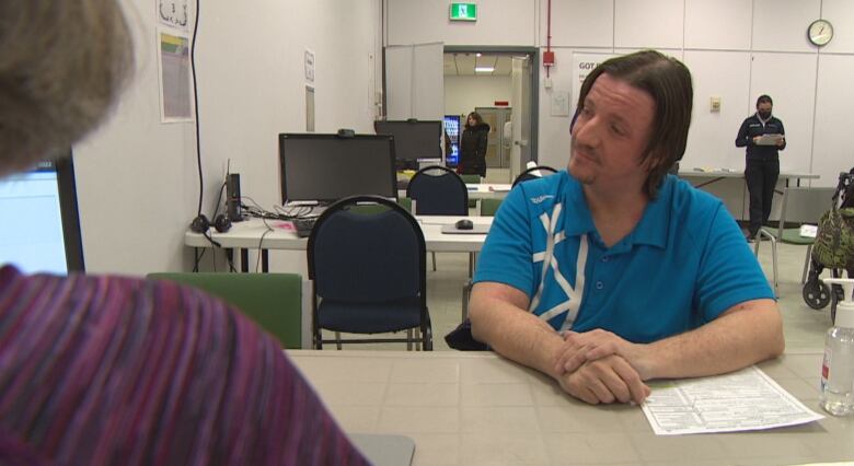 Man with medium-long hair in a blue t-shirt sits at a table facing a woman at a laptop computer who is asking him questions about his tax forms.