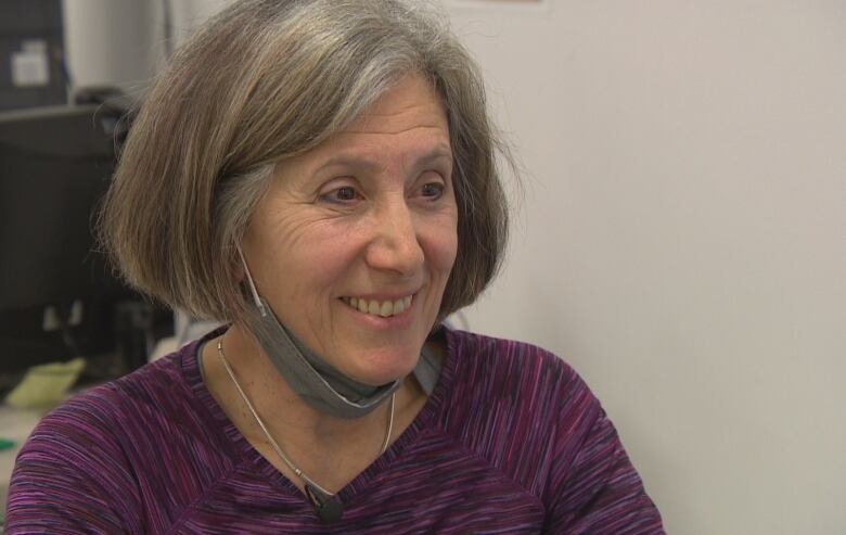 A woman with a grey bob hairstyle and sporty purple shirt smiles while sitting at her desk, her medical mask lowered for the photo.