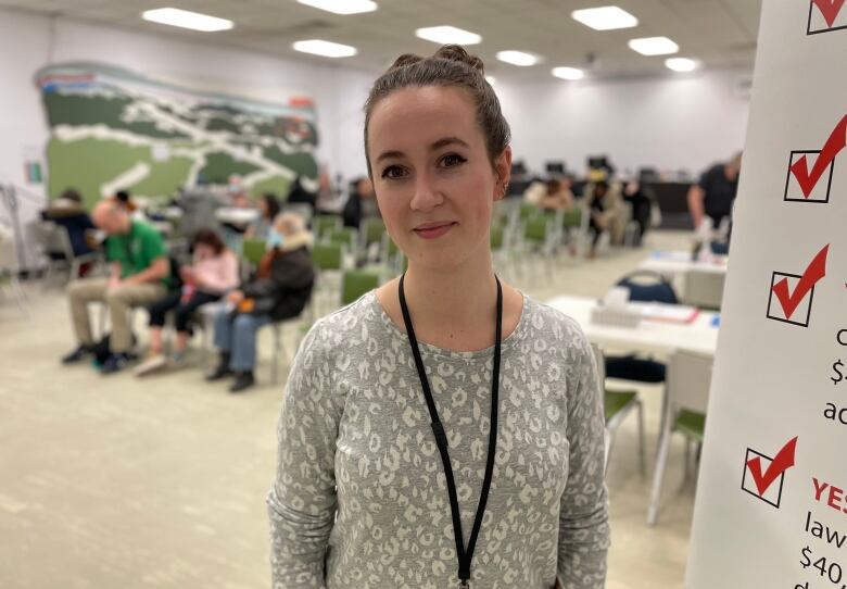 Young woman wearing a grey patterned shirt with her brown hair put into a bun stands in front of a busy waiting room filled with people who've registered for help with their taxes.