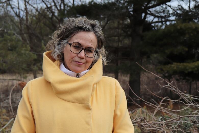 A woman in glasses and a yellow jacket stands outside, looking at the camera. 