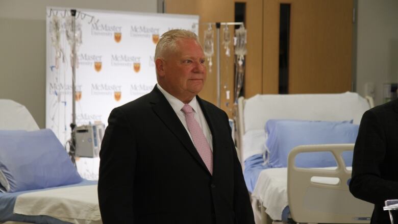 A person in a suit stands in front of hospital beds and IV bags.