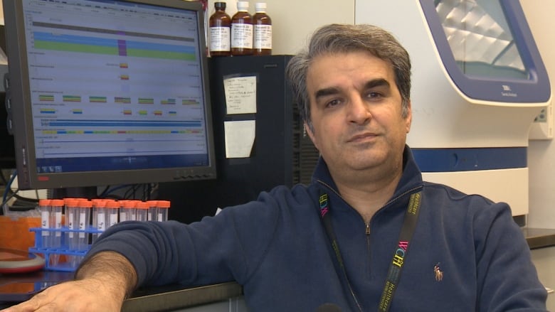 A man sits in front of a computer and some test tubes in a lab, looking at the camera