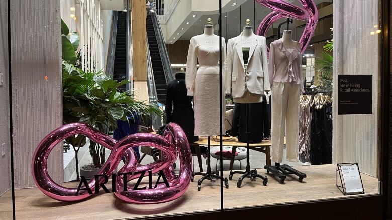 A pink, curved sculpture is pictured inside a window display for a store inside a mall. Women's clothing is pictured on nearby mannequins.